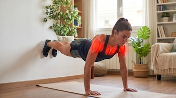 Mujer con coleta haciendo flexiones contra una pared blanca. Sus pies están apoyados en la pared y sus manos en una esterilla. Músculos del torso resaltados en rojo.