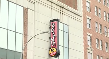 Un letrero vertical rojo con la marca ShopRite en la fachada de un edificio beige junto a un edificio residencial de ladrillo rojo