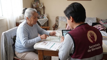 Mujer adulta de cabello canoso sentada en mesa con documentos, señalando una identificación. Joven con chaleco de Bienestar usa tableta digital.