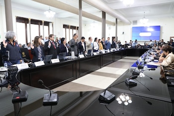 Vista interior de una sala de conferencias con un grupo de personas de pie con la mano derecha levantada ante una mesa, mientras otras están sentadas