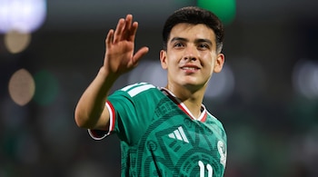 Un hombre vestido con la camiseta verde de la selección de México levanta la mano derecha y sonríe en un estadio.