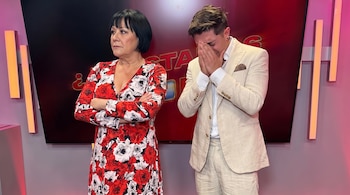 Mujer de cabello oscuro y vestido floral rojo y blanco con brazos cruzados junto a hombre con traje claro que se cubre el rostro con las manos.