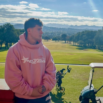 Tomás Mazza viste una sudadera rosa en un campo de golf con un carro de golf a la derecha y colinas al fondo