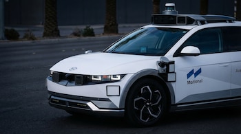 Frente y costado de un vehículo autónomo blanco de Motional con sensores en el capó, laterales y techo. Ruedas negras y plateadas visibles. Palmeiras al fondo.