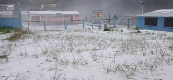 Un terreno cubierto de nieve con hierba, una valla metálica con cancha de baloncesto y edificios al fondo bajo la niebla