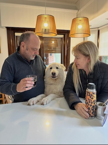 Un hombre y una mujer sentados junto a un perro blanco frente a una mesa con un vaso, un termo y un mate