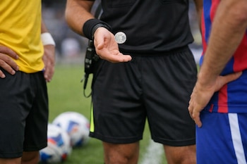 Un árbitro lanza una moneda al aire con la palma abierta, flanqueado por dos jugadores de fútbol en una cancha de césped con balones de fondo.