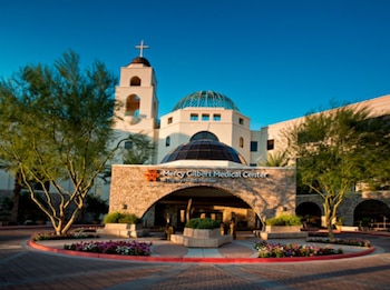 Entrada principal del Mercy Gilbert Medical Center con edificio blanco, cúpula de cristal, torre con cruz, arco de piedra, árboles y flores bajo cielo azul