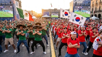 Una multitud de aficionados mexicanos y surcoreanos baila el 'Gangnam Style' en un Fan Fest. Muestran banderas de ambos países y pantallas gigantes con un partido al atardecer.