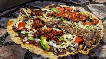 Tortilla tostada grande cubierta con frijoles, queso, chorizo, tiras de tasajo, jitomate y aguacate sobre una mesa de piedra.