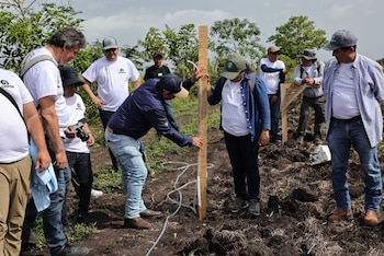 Grupo de personas en un campo de tierra con árboles y arbustos. Dos personas miden el suelo con una estaca de madera y un tubo translúcido