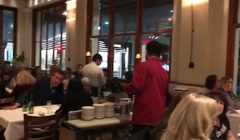 Salón de un restaurante con clientes sentados a las mesas, camareros con uniformes de servicio y ventanales con vista a la calle por la noche