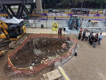 Una excavadora junto a un foso acordonado, una cabina de teleférico y varias personas reunidas en la zona de obra