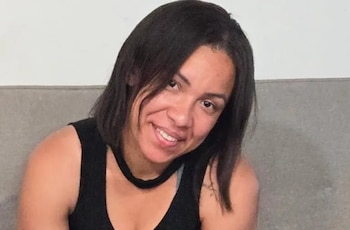 Una mujer sonriente de cabello oscuro y camiseta negra posa sentada frente a un respaldo gris