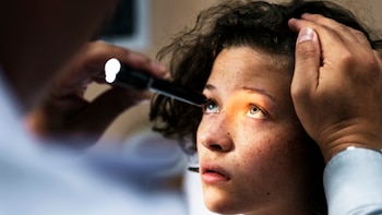 Primer plano de una persona joven con pecas, cuyo ojo está siendo iluminado por una linterna sujeta por una mano, mientras otra mano apoya la frente