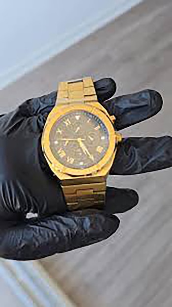 Una mano cubierta con un guante negro sostiene un reloj de pulsera dorado con esfera oscura