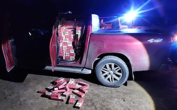 Una camioneta roja de noche con la puerta abierta llena de paquetes rojos de cigarrillos y varios cartones apilados en el suelo de tierra