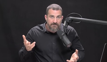 Hombre con barba y camisa oscura habla ante un micrófono con el brazo izquierdo levantado. El fondo es completamente negro