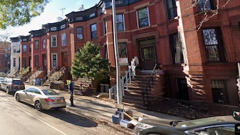 Vista de una calle con una hilera de edificios de ladrillo de varios pisos, escaleras frontales, acera con árboles, SUV y coches estacionados