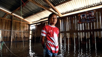 Uma menina parada na água de uma casa de madeira inundada com telhado de ferro; Vestindo uma camiseta vermelha e branca e shorts.