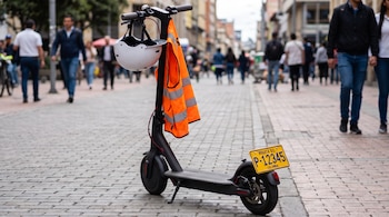 Una patineta eléctrica negra con casco blanco y chaleco reflectivo naranja colgados del manubrio. Incluye placa amarilla de Bogotá D.C., Colombia. Fondo de calle peatonal con gente.