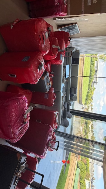 Múltiples maletas y bultos voluminosos envueltos en plástico rojo apilados en el suelo de una sala moderna con grandes ventanales y vista a un jardín