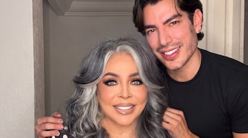 Retrato de una mujer con cabello gris ondulado y un hombre con cabello oscuro. Ambos sonríen, el hombre apoya una mano en el hombro de la mujer.