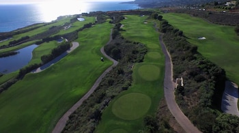 Vista aérea de un campo de golf con césped verde, lagos, caminos pavimentados, trampas de arena, vegetación, y el océano bajo un cielo soleado