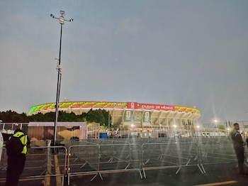 Vista exterior del Estadio Ciudad de México al anochecer, con su fachada iluminada en verde y rojo, barreras metálicas y una persona de seguridad en primer plano