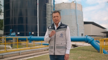 Un hombre con chaleco que gesticula con la mano frente a dos grandes tanques industriales y tuberías de color azul.