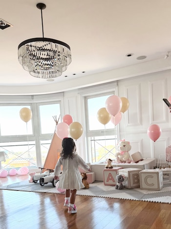 Una niña con vestido blanco camina de espaldas en una sala decorada con globos rosados y beige, juguetes de peluche y cubos con letras