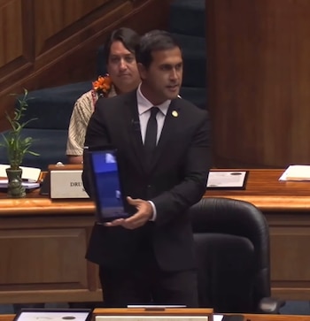 Hombre en traje negro habla en podio, sostiene una tableta con pantalla azul. Otro hombre con camisa estampada está sentado detrás. Fondo de paneles de madera