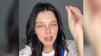 Una mujer con cabello oscuro y ojos azules posa de frente, con su mano derecha elevada y los dedos unidos. Se observa un objeto azul en la parte inferior.