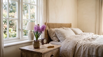 Planta de jacinto en maceta, libro y reloj sobre mesa de noche de madera al lado de una cama con cojines y una ventana.