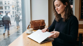 Una mujer con suéter oscuro cierra una laptop plateada en una mesa de madera. Junto a ella, una taza de café y un bolso. Detrás, una calle urbana.