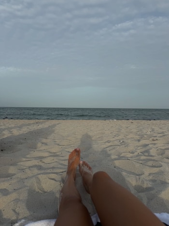Pies y piernas de una persona recostada sobre la arena de una playa, con el mar y un cielo nublado visible al fondo