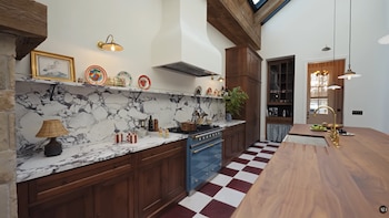 Vista interior de una cocina con techo alto y vigas, gabinetes de madera, estufa azul, mármol en el salpicadero, y un suelo de baldosas a cuadros