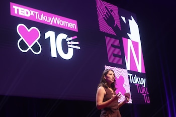 Una mujer con ropa oscura habla en un escenario frente a una pantalla morada. En la pantalla se leen 'TEDxTukuyWomen', '10 años de ideas valientes' y diseños gráficos