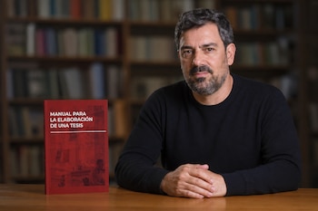 Un hombre con barba y suéter negro sentado en una mesa de madera junto a un libro rojo y estanterías con libros de fondo.