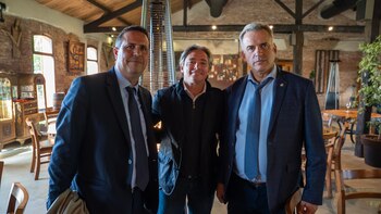 Tres hombres con traje y vestimenta formal posan de pie en el interior de un salón con paredes de ladrillo visto y vigas de madera