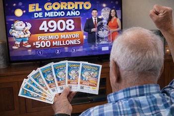 Hombre de espalda sostiene boletos de lotería frente a una televisión que muestra un sorteo con dos presentadores, el número 4908 y un premio mayor.