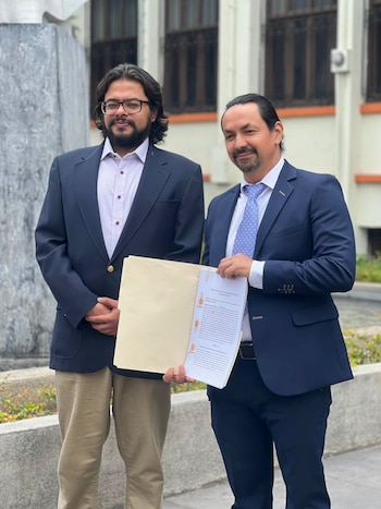 Dos hombres de pie al aire libre. Uno lleva saco azul y pantalón claro. El otro, traje azul y corbata, sostiene documentos con texto impreso