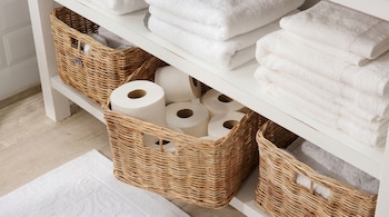 Estantería blanca con toallas blancas dobladas en la parte superior y en cestas de mimbre. Las cestas también contienen rollos de papel higiénico. Un tapete blanco se ve en el suelo.