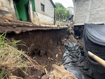 Gran deslizamiento de tierra entre dos casas; el suelo erosionado expone cimientos. Un hombre mira el área afectada; bolsas de Quikrete Concrete Mix visibles