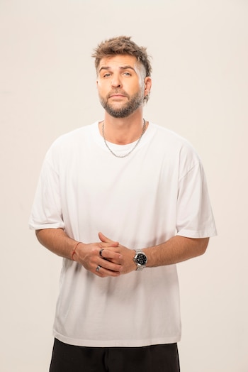 Un hombre caucásico con barba y cabello castaño corto, vistiendo una camiseta blanca, pantalón oscuro, cadena, anillos y reloj, posa con manos entrelazadas sobre fondo beige