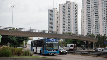Un colectivo azul y blanco de la línea 74 en una avenida urbana bajo un puente, con edificios altos y vegetación en el fondo, bajo cielo nublado