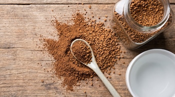 Café instantáneo en polvo y en gránulos, una cuchara con café, un frasco de vidrio lleno de gránulos y una taza blanca vacía sobre una mesa de madera.