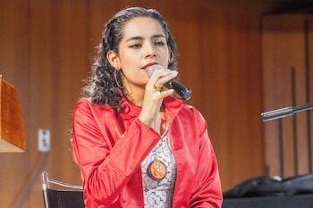 Una mujer de cabello oscuro sostiene un micrófono negro mientras viste una chaqueta roja y un collar con dije