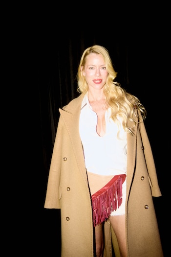 Nicole Neumann frente a un fondo negro con un abrigo color camel, una camisa blanca y una falda con flecos rojos
