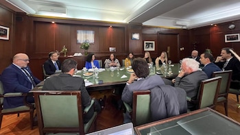 Una sala de conferencias con once personas sentadas alrededor de una mesa de madera. En la mesa hay vasos, platos, documentos y un ordenador